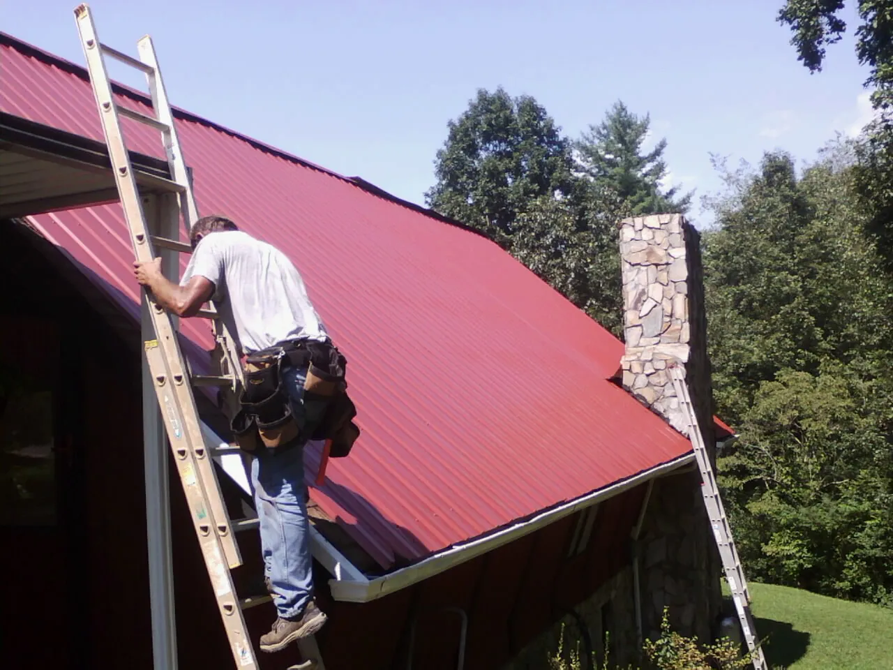 Roofer on a ladder beside a red metal roof and stone chimney — Soffit & Fascia Repair by Roofing Brothers in Granger